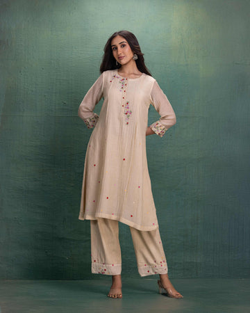 Woman wearing a cream-colored embroidered kurta set with straight pants, styled with minimal jewelry and heels, posing against a green textured background.
